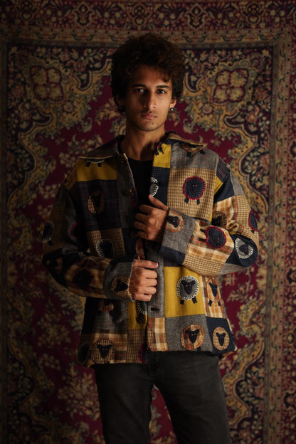 A person standing in front of a patterned backdrop wearing a multicolored patchwork shirt jacket with sheep applique.