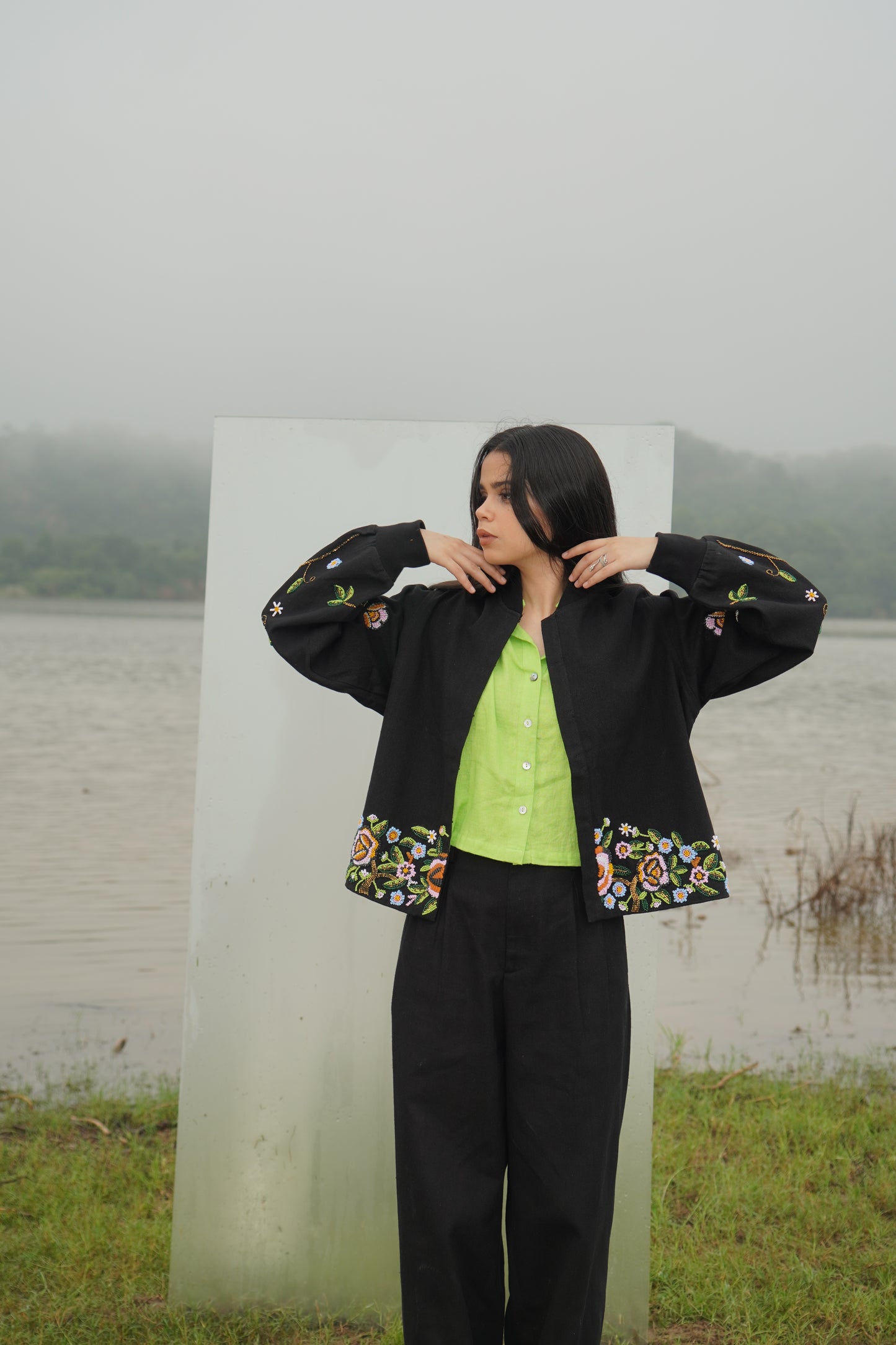 A person wearing a black unisex jacket with floral embellishments and a green top, standing near a transparent panel with a natural background.